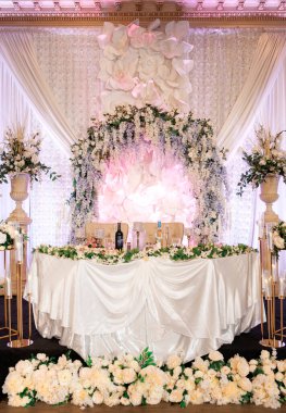 Beyaz dekorlu bir restoranda gelin ve damat için şenlikli bir düğün masası. Çiçek aranjmanlı bir düğün masası ve beyaz çiçeklerden yapılmış bir kemer. Düğün yemeği için zarif beyaz tema