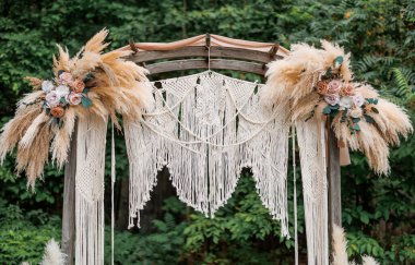 Düğün kemeri çiçekler ve makrome ile süslenmiş. Açık havada bir düğün töreni için kırsal bir ahşap kemerin altında asılı bir Macrame. Kuru ot ve güllerle süslü çiçek kompozisyonları