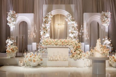 Gösterişli bir düğün resepsiyonu, bej renkli perdeler ve zengin çiçeklerle süslenmiş. Romantik bir ortam için beyaz kemerler ve mumlarla süslenmiş güzel çiçekli bir düğün sahnesi.