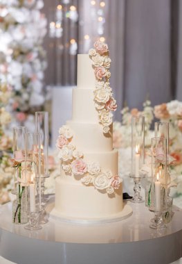 Beyaz ve pembe krem güllerle süslenmiş çok katlı bir düğün pastası. Düğün ziyafetinde çiçeklerle süslenmiş zarif bir düğün pastası ve romantik bir ortam için mum yakmak.