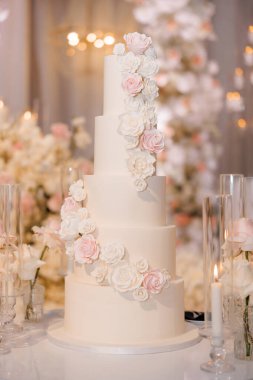 Beyaz ve pembe krem güllerle süslenmiş güzel katlı bir düğün pastası. Arka planda çiçeklerle süslenmiş bir restoranda şık bir düğün pastası. Beyaz kremalı tatil pastası.