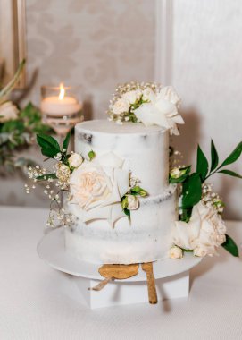 Beyaz güllerle süslenmiş beyaz bir düğün pastası ziyafette masanın üzerinde duruyor. Vanilya kremalı, taze çiçeklerle süslenmiş iki katlı küçük bir tatil pastası. Misafirler için tatlı ikram.