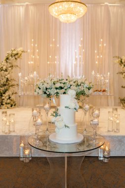 Taze beyaz güllü nefis kremalı düğün pastası. Üç katlı beyaz düğün pastası romantik dekorasyonların ortasında çiçekler ve mumlarla. Lüks bir restoranın içinde vanilyalı kek.