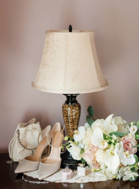 Gelinin düğün aksesuarları masa lambasının yakınındaki odasında. Gelinin buketi beyaz çiçekler, topuklu ayakkabılar, el çantası, alyans ve düğün günü için hazırlanmış parfüm.