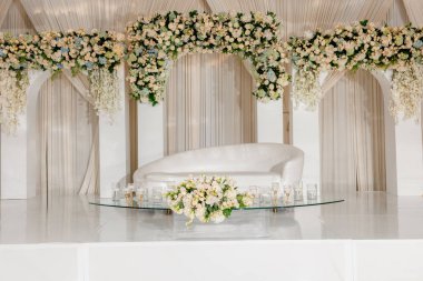 Beyaz kanepesi ve çiçekleri olan zarif bir düğün sahnesi. Yeni evliler için yapılan düğün töreninin sahnesi gür çiçek kemerleri ve beyaz ve krem rengi perdelerle süslenmiştir. Düğün geçmişleri