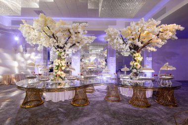 Beyaz çiçek açan ağaçlar ve kristal avizelerle süslü bir düğün resepsiyonu. Büyük cam bir masada gümüş tabaklar ve kapalı bir düğün yemeği için romantik bir ortam.