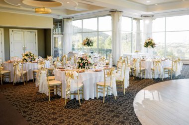 Düğün için süslenmiş masaları olan bir salon. Çatal bıçak ve çiçeklerle servis edilen şenlikli masalar ve sandalyeler. Düğün resepsiyonu ya da büyük pencereli büyük bir salonda şenlik.