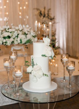 Beyaz vanilyalı kremayla kaplı üç katlı güzel düğün pastası. Mumların ortasındaki bir ziyafette taze güllerle süslenmiş beyaz bir pasta restoranda romantik bir ortam yaratıyor.