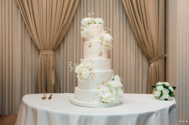 Restoranda çok katmanlı beyaz bir düğün pastası. İçerde taze orkideler ve güllerle süslenmiş kremalı pasta. Bütün düğün pastası, yeni evliler için hazırlanmış bir masada tek başına duruyor.