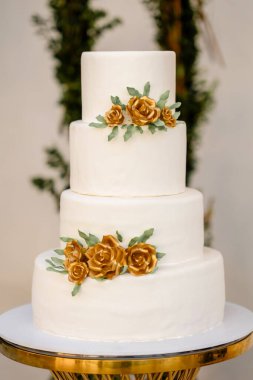 Altın güllerle süslenmiş beyaz düğün pastası yakın plan masa üzerinde duruyor. Dört katlı düğün pastası, beyaz krema ve şeker gülleriyle kaplı. Düğün salonundaki yuvarlak masada duruyor.