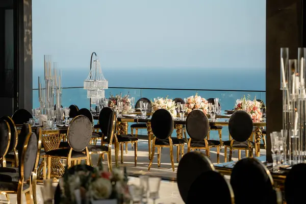 Düğünde misafirler için terasta masa servisi yaptım. Kadife siyah sandalyeler, kristal lambalar ve çiçeklerle lüks bir şölen. Zarifçe okyanus manzaralı yemek masaları kurun.