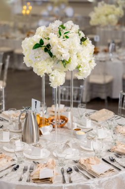 Merkezinde uzun beyaz çiçeklerle süslenmiş zarif bir düğün masası bayramın zarif ve romantik bir atmosferini yaratır. Çatal bıçak takımı ve hediye kutularıyla dolu küçük yuvarlak bir masa.