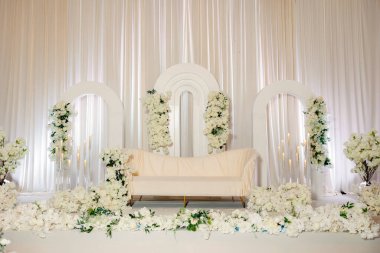 Beyazlı gelin ve damat için büyük bir düğün sahnesi. Beyaz çiçekleri, kemerleri, mumları ve kanepesi olan zarif bir düğün sahnesi düğün töreni ve fotoğrafçılık için mükemmel bir arka plan.