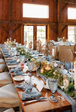 Zarif mavi cam eşyalar, mumlar, yeşillik ve çiçek düzenlemeleri ile taşralı düğün masası ahşap ahır mekanında sıcak ve davetkar bir ortam yaratıyor.