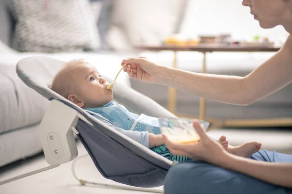 母亲用勺子在婴儿椅上用果冻喂男婴 婴儿固体食物介绍概念 — 图库照片