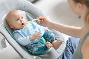 Anne, bebeğini bebek koltuğunda meyve püresiyle besliyor. Bebek katı gıda tanıtım konsepti