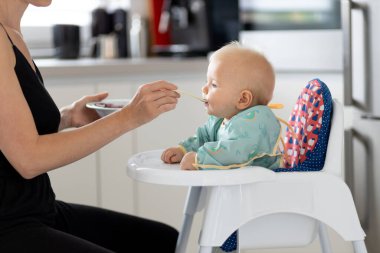 Anne, bebeğini evde meyveli püre ile bebek koltuğunda besliyor. Bebek katı gıda tanıtım konsepti.