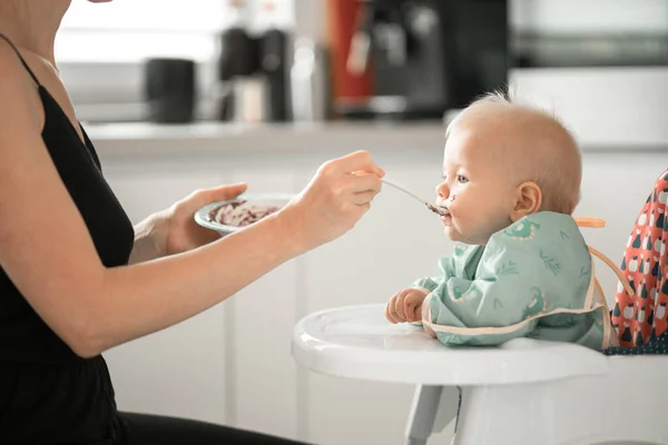 母親のスプーンは赤ん坊の椅子の彼女の赤ん坊の男の子の子供に家の台所の果物のピューレを与える 赤ちゃん固形食導入コンセプト ストック写真