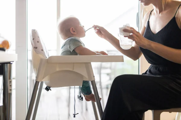 妈妈的勺子在家里的婴儿椅子上喂她的男婴 在厨房里放着水果泥 婴儿固体食物介绍概念 — 图库照片