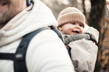 Yavru oğlunu taşıyan sportif baba sonbahar ormanlarında yürüyüş yaparken sırt çantasında tulum ve şapka giyiyor.
