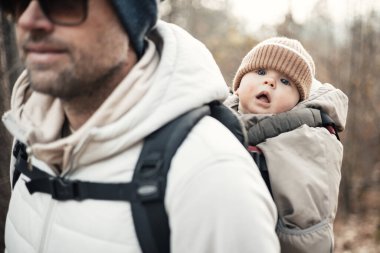 Yavru oğlunu taşıyan sportif baba sonbahar ormanlarında yürüyüş yaparken sırt çantasında tulum ve şapka giyiyor.