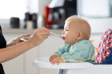 Anne, bebeğini evde meyveli püre ile bebek koltuğunda besliyor. Bebek katı gıda tanıtım konsepti