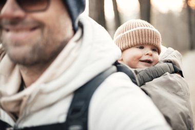 Yavru oğlunu taşıyan sportif baba sonbahar ormanlarında yürüyüş yaparken sırt çantasında tulum ve şapka giyiyor.