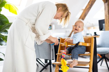 Happy infant sitting and playing with his toy in traditional scandinavian designer wooden high chair in modern bright atic home suppervised by his mother