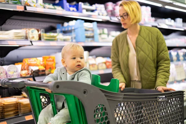 Casualy dressed mother choosing canned products in department of supermarket grocery store with her infant baby boy child in stroller