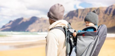 Young father carrying his infant baby boy sun in backpack on windy sandy beach of Famara, Lanzarote island, Spain. Family travel and winter vacation concept. Parenting, fatherhood, fathers day