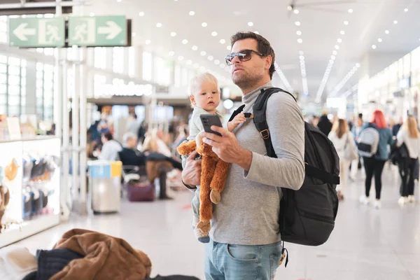 父は子供と一緒に旅行し 空港ターミナルで幼児の男の子を抱えて フライトスケジュールをチェックし 飛行機に乗るのを待っています 子供と一緒に旅行するコンセプト — ストック写真