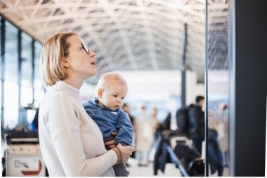 Anne çocuğuyla seyahat ediyor, bebeğini havaalanı terminalinde tutuyor uçuş saatlerini kontrol ediyor, uçağa binmeyi bekliyor. Çocuk konseptiyle seyahat