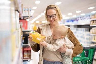 Caucasian mother shopping with her infant baby boy child choosing products in department of supermarket grocery store