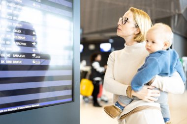 Anne çocuğuyla seyahat ediyor, bebeğini havaalanı terminalinde tutuyor uçuş saatlerini kontrol ediyor, uçağa binmeyi bekliyor. Çocuk konseptiyle seyahat