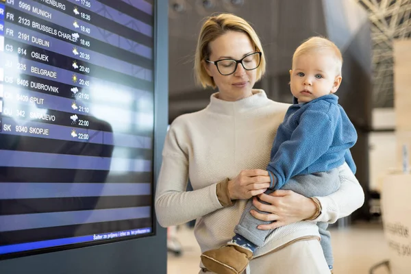 母亲带着孩子旅行 在机场候机楼抱着他的男婴 检查航班时刻表 等待登机 带孩子旅行的概念 — 图库照片