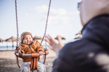 Anne, neşeli erkek bebeğini İspanya 'nın Malaga şehrinde güneşli bir kış günü kumsalda salıncakta sallıyor.