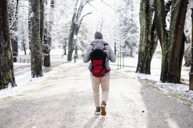 Yavru oğlunu sırtında taşıyan sportif bir babanın kışın şehir parkında yürüdüğü sırt çantasındaki tulum ve şapkasını..