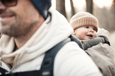 Yavru oğlunu taşıyan sportif baba sonbahar ormanlarında yürüyüş yaparken sırt çantasında tulum ve şapka giyiyor.