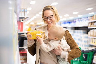 Caucasian mother shopping with her infant baby boy child choosing products in department of supermarket grocery store