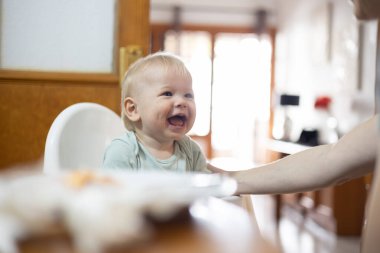 Sevimli, mutlu, erkek bebek bebek. Mutfaktaki yemek masasında yüksek sandalyede otururken gülümseyen çocuk. Annesi tarafından kaşıkla besleniyor..