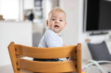 Geleneksel İskandinav tasarımcı tahta sandalyede oturan ve modern aydınlık evde yüksek sesle gülen mutlu bir bebek. Sevimli bebek gülüşü