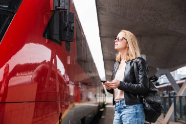 Kot pantolon, gömlek ve deri ceketli, çanta ve güneş gözlüğü takan genç sarışın kadın tren istasyonunda kırmızı hız trenine biniyor. Seyahat ve ulaşım