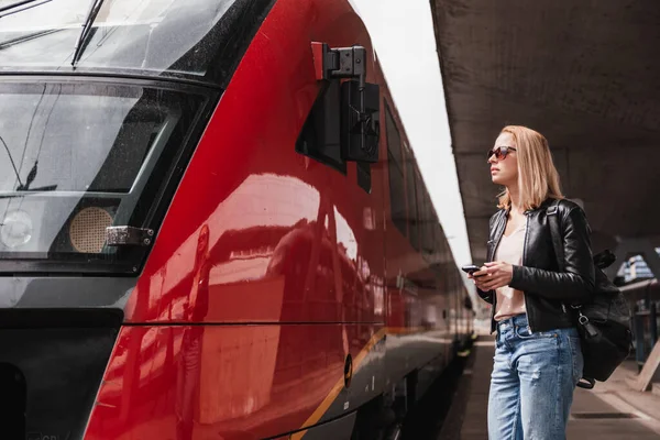 stock image Young blond woman in jeans, shirt and leather jacket wearing bag and sunglass, embarking red modern speed train on train station platform. Travel and transportation