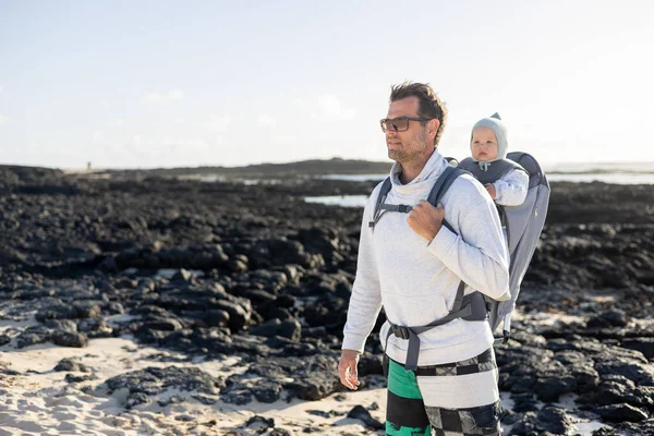 スペインのランサローテ島の黒い岩の火山のビーチでリュックに彼の幼児の男の子の息子を運ぶ若い父親 家族旅行と冬の休暇のコンセプト — ストック写真