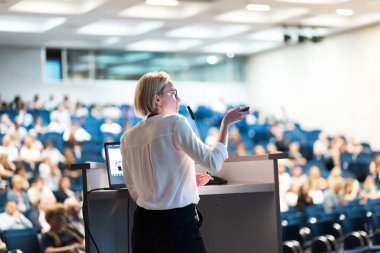 Female speaker giving a talk on corporate business conference. Unrecognizable people in audience at conference hall. Business and Entrepreneurship event clipart