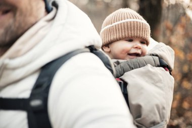 Yavru oğlunu taşıyan sportif baba sonbahar ormanlarında yürüyüş yaparken sırt çantasında tulum ve şapka giyiyor.