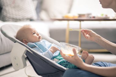 Anne, bebeğini bebek koltuğunda meyve püresiyle besliyor. Bebek katı gıda tanıtım konsepti