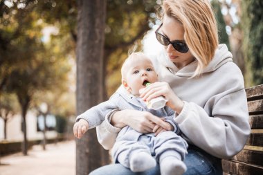 Anne şehir parkında bankta oturuyor, bebeğini kucağında tutuyor ve güneşli bir bahar gününde ona bebek maması yediriyor..