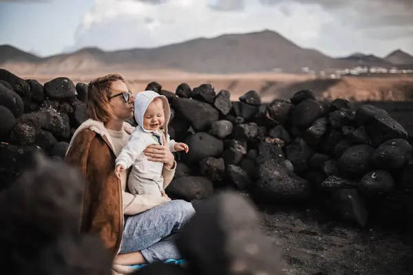Mother enjoying winter vacations playing with his infant baby boy son on black sandy volcanic beach of Janubio on Lanzarote island, Spain on windy overcast day. Family travel vacations concept
