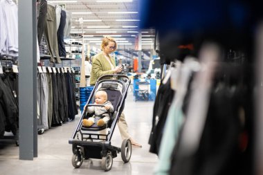 Casualy dressed mother choosing sporty shoes and clothes products in sports department of supermarket store with her infant baby boy child in stroller